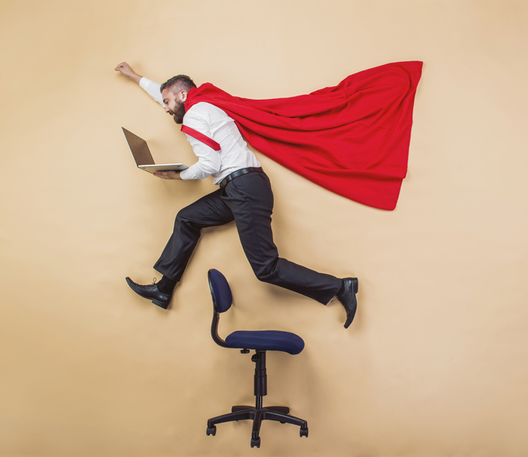 man with Superman cape leaps over steno chair with his laptop open in his hands
